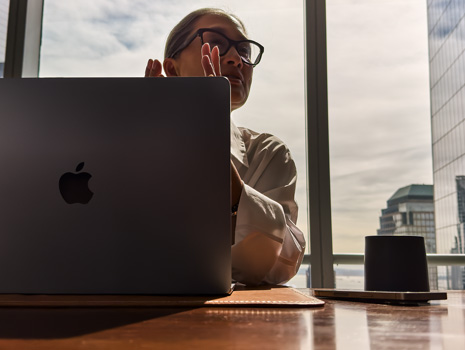 Kobieta siedząca przy stole w wieżowcu. Przez duże okno za nią widać inne budynki. Przed nią stoi otwarty MacBook.