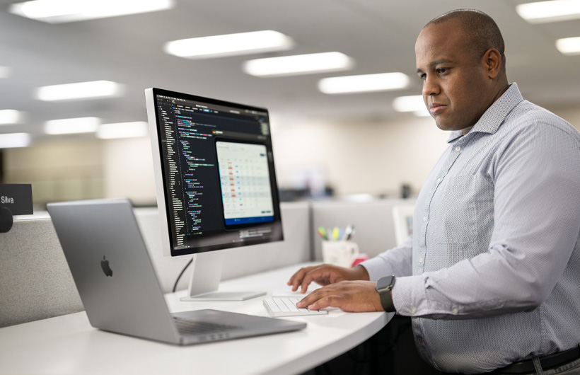 Un développeur debout à son bureau utilise un MacBook et un Mac affichant des lignes de code et les informations d’une app